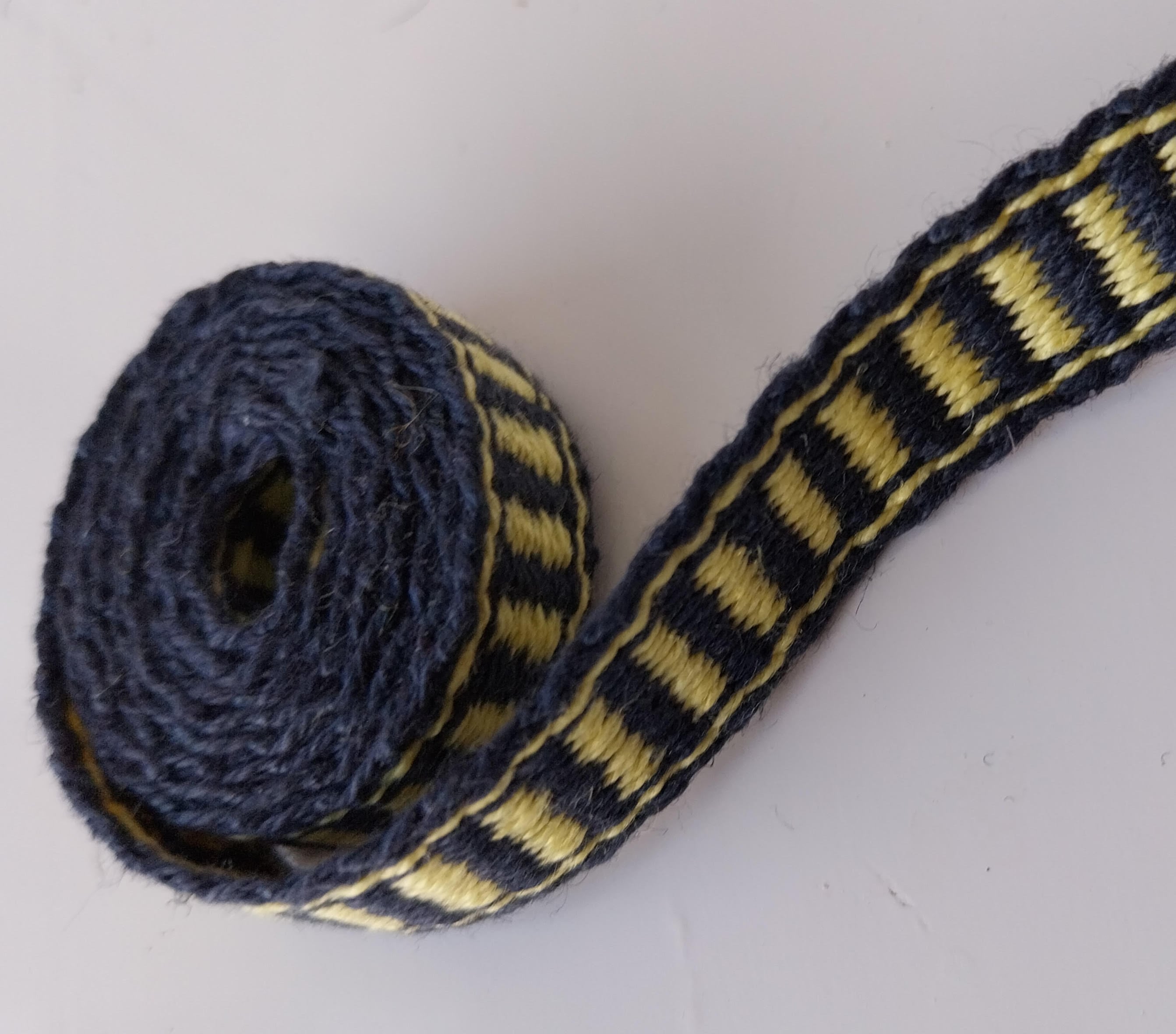 Tværstribet bånd med 3 langsgående striber som kant. det samme som på billedet med sko og jakke.
Bomuld 16/2. 31 tråde. 1,2 cm bredt. Enderne har fået en takling, og er dyppet i fortyndet snedkerlim.